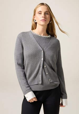 Grauer gerippter Cardigan mit V-Ausschnitt, ausgestattet mit einer Knopfverschluss, Perlenhalsketten-Detail und weißen Bündchen, die aus einem dunklen Outfit darunter herausragen.