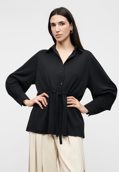 Γυναίκα με μακριά σκούρα μαλλιά φοράει μαύρο πουκάμισο με μακριά μανίκια και κουμπιά, δεμένο στη μέση, και μπεζ πλισέ φαρδιά παντελόνια, ποζάροντας με τα χέρια στη μέση.