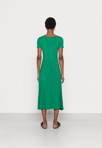 Vestido verde, de manga curta, até ao joelho, com corpete ajustado e saia fluida. Feito de um tecido suave com ligeira elasticidade. Sandálias rasas.