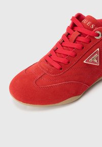 Piros velúr magas szárú tornacipő texturált kontúrokkal, hozzáillő fűzőkkel, és egy arany logó díszítéssel az oldalon, szürke háttér előtt.