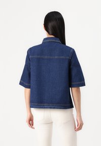 Kratka modra denim jakna s kratkimi rokavi, ovratnikom, vidnimi šivi in ravnim krojem. Nosi se čez svetlo obarvane hlače.