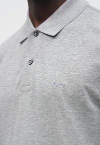 Szara koszulka polo z teksturowanej tkaniny, z kołnierzykiem, dwoma guzikami oraz małym, haftowanym logo "BOSS" na piersi.