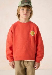 Sweatshirt orange avec un graphique rond "GOOD VIBES" en bleu clair. Le tissu semble doux, avec une coupe décontractée et des poignets côtelés.