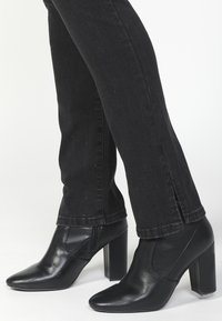 Botas de tobillo de cuero negro con punta afilada, tacón alto y cuadrado, y detalle de cremallera lateral, usadas con jeans negros ajustados.