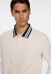 Beige polo skjorta med en randig svart och vit krage. Har en två-knappsförslutning och en liten broderad logotyp på bröstet.