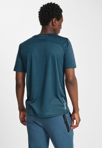 T-shirt de sport en vert foncé à manches courtes, texture lisse et un subtil détail de logo dans le dos, porté avec un pantalon bleu assorti doté de poches zippées.