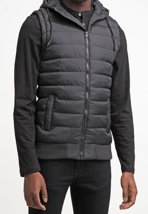 Mężczyzna w czarnej pikowanej kamizelce bez rękawów z kapturem, na czarnej bluzce z długim rękawem i czarnych spodniach, stojący z rękami swobodnie opuszczonymi wzdłuż ciała.