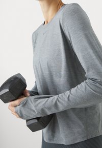 Harmaa pitkähiha urheilupaita, jossa on sileä pinta, pyöreä kaulus ja istuvat hihansuut, pitelee mustaa kuusikulmaista käsipainoa.