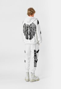 Sudadera blanca con gráficos de alas negras en la espalda, combinada con joggers blancos que presentan diseños similares y detalles en texto, usados con botas plateadas.