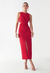 Red, sleeveless midi dress with a twist front and side cutouts, featuring a thigh-high slit. Smooth texture, complemented by metallic accessories.