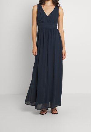 Femme portant une robe longue sans manches bleu marine avec col en V et taille froncée, et des sandales noires à brides, debout devant un fond uni.