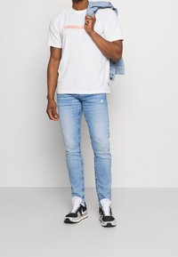 Vit bomullströja med "UNAVAILABLE" i rosa bokstäver, ljusblå slim-fit-jeans och svart-vita sneakers. En denimjacka hängande över axeln.