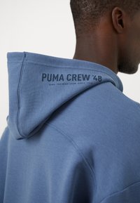 Lyseblå hættetrøje lavet af blødt stof. Har en syet tekstur og et sort trykt mærke på ryggen med teksten "PUMA CREW '48."