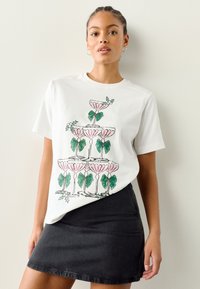 Maglietta bianca in cotone con grafica di bicchieri di champagne rosa con fiocchi verdi e foglie. Abbinata a una gonna di denim scuro.