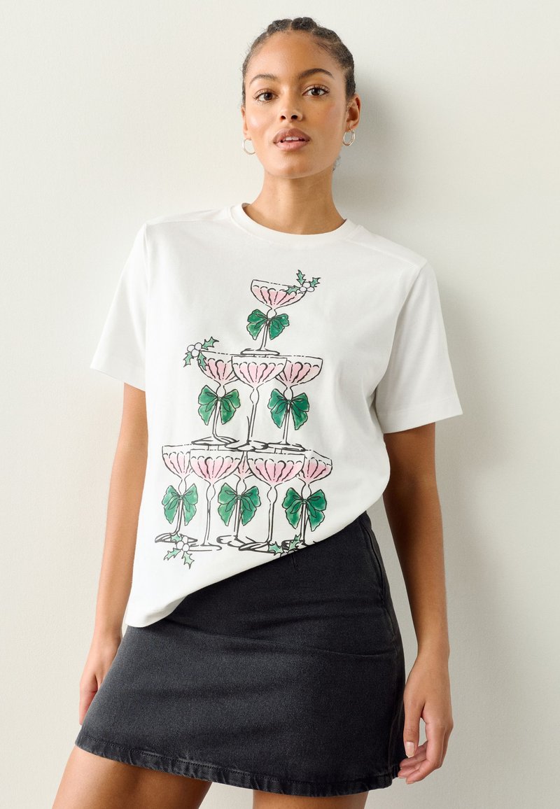 Maglietta bianca in cotone con grafica di bicchieri di champagne rosa con fiocchi verdi e foglie. Abbinata a una gonna di denim scuro.