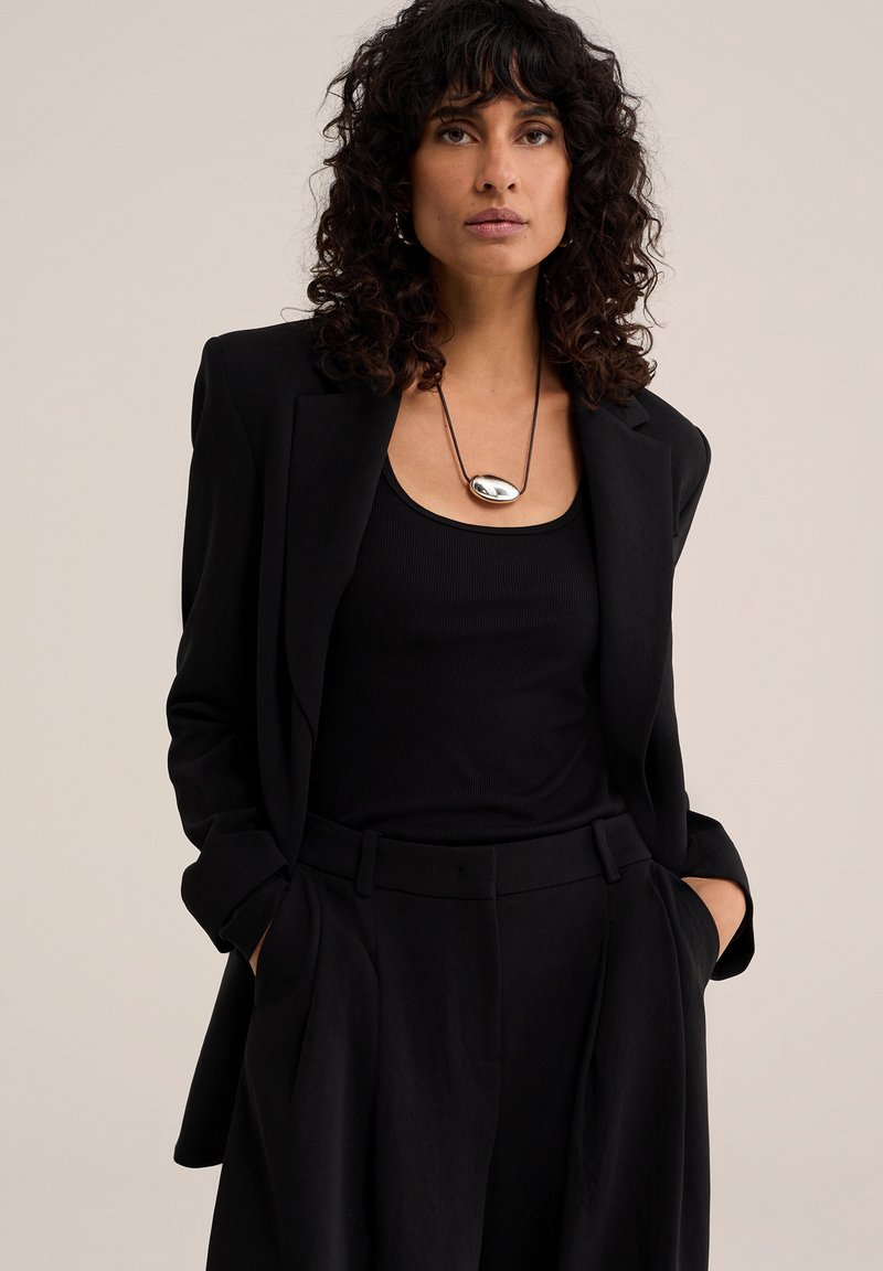 Femme aux cheveux bouclés foncés porte un blazer et un pantalon noirs, un haut noir et un collier pendentif ovale en argent, debout les mains dans les poches.
