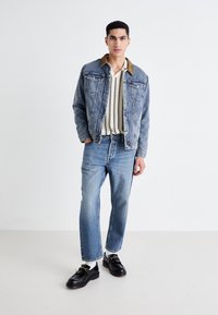 Denim jakk musta pruun krae, vertikaalsete triipudega särk, heledad sinised teksad ja mustad loafersid kuldsete aktsentidega. Mudel seisab lihtsas taustas.