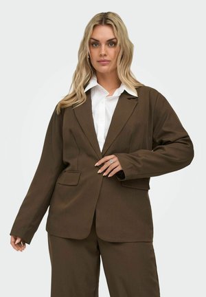 Lyshåret kvinde iført en brun oversized jakkesætjakke og matchende bukser over en hvid skjorte med krave, poserer med den ene hånd på jakken.