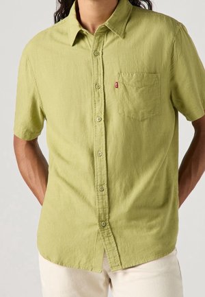 Homme portant une chemise à manches courtes vert clair avec une poche avant et un pantalon beige, debout les mains derrière le dos.