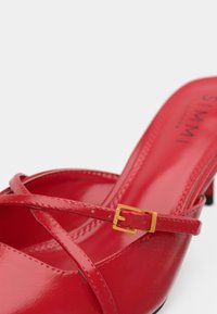Červená lakovaná vysoká podpätková topánka s prekríženými remienkami a malou zlatou sponou na bielom pozadí.