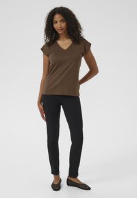 T-shirt marrone a V con maniche corte, vestibilità slim, abbinato a pantaloni slim neri. Il modello indossa ballerine nere in rete. Sfondo semplice.