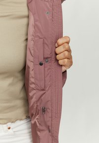 Helle rosa Nylonjacke mit einem Reißverschlusstasche und Druckknopfschlüssen, mit sichtbaren Stichdetails entlang der Öffnung und am Ärmel.