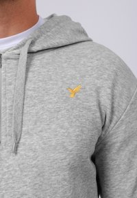 Man wearing light grey zip-up hoodie with a small yellow embroidered hummingbird on the left chest area.