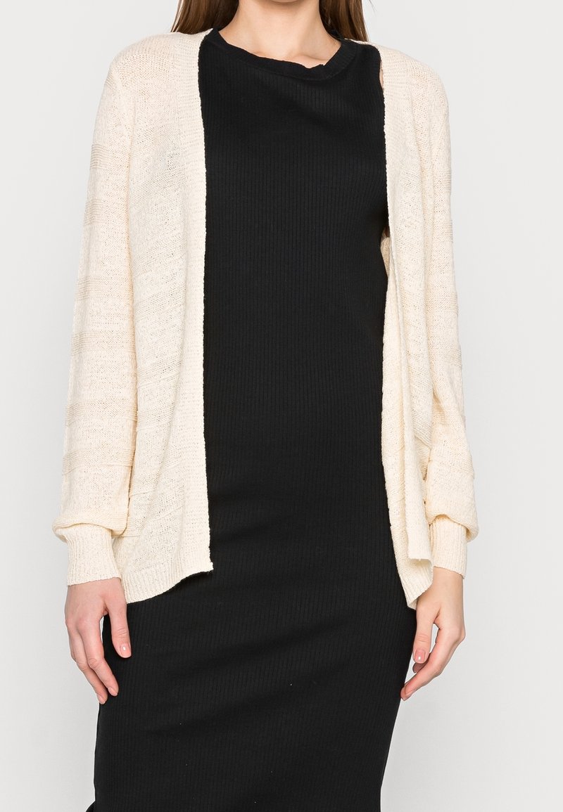 Ljusbeige kofta med öppen framtill, ribbat mönster, vida ärmar och avslappnad passform, bärs över en svart ribbad klänning.