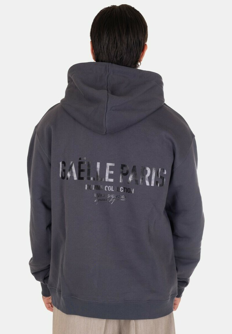 Felpa grigia scura con cappuccio presenta un grande logo "GAËLLE PARIS" in argento riflettente sul retro, con un cappuccio spazioso e polsini a costine.