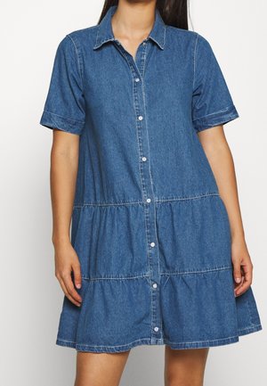 Mujer vistiendo un vestido informal de mezclilla azul con mangas cortas, botones en la parte delantera y detalles de falda en volantes.