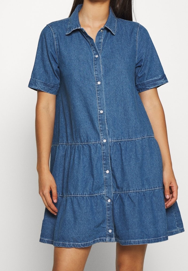 Femme portant une robe casual en denim bleu à manches courtes avec des boutons sur le devant et des détails de jupe à étages.
