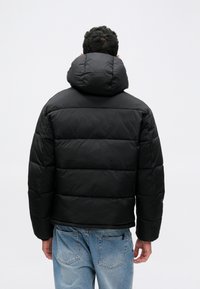 Bărbat îmbrăcat într-o jachetă neagră căptușită cu glugă, întors cu spatele, purtând blugi albaștri deschis, pe un fundal alb simplu.