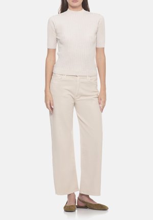 Donna in piedi che indossa una maglietta corta a costine color crema, pantaloni beige a gamba larga e scarpe basse verde oliva su sfondo bianco.