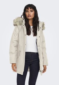 Femme portant une parka beige avec une capuche en fourrure, un chemisier blanc et un jean noir, se tenant devant un fond clair uni.