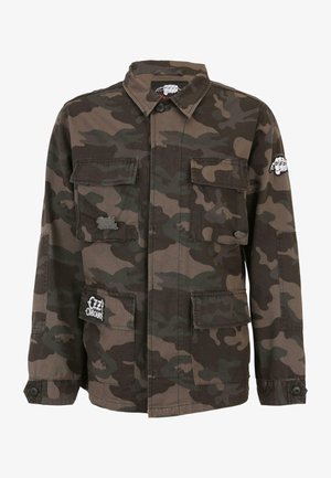 Kurtka w kamuflażu z mieszanki bawełny, z dwoma kieszeniami na piersi, zapięciem na guziki i haftowanymi akcentami. Dominujące kolory to ciemna zieleń i brąz.