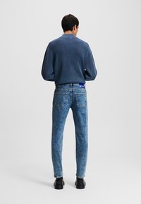 Un pull bleu côtelé avec un col, associé à un pantalon en jean bleu délavé. Le jean a un design classique à cinq poches et est porté avec des chaussures noires.