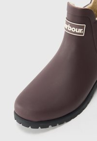 Sötétbarna vízálló csizma fekete barázdás talppal és világosbarna bolyhos béléssel, a bokához közel pedig egy fehér kerettel ellátott "Barbour" logó található.