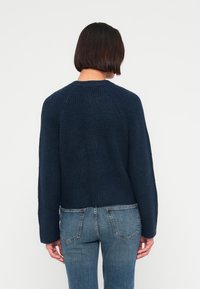 Μπλε σκούρο πλεκτό πουλόβερ με ανάγλυφη υφή, στρογγυλή λαιμόκοψη και μακριά μανίκια. Φορεμένο πάνω από ανοιχτόχρωμα τζιν, δείχνοντας casual εφαρμογή.