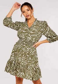 Olivenfarbenes Kleid mit weißen Zebra-Streifen, langen Ärmeln, Knopfleiste und gestuftem Rüschensaum. Hergestellt aus leichtem Stoff.