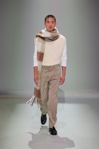 Modellen har en cremefarvet sweater på med en beige vest, lysebeige bukser og sorte sko, suppleret med et stort tørklæde med ternet mønster i bløde farver.
