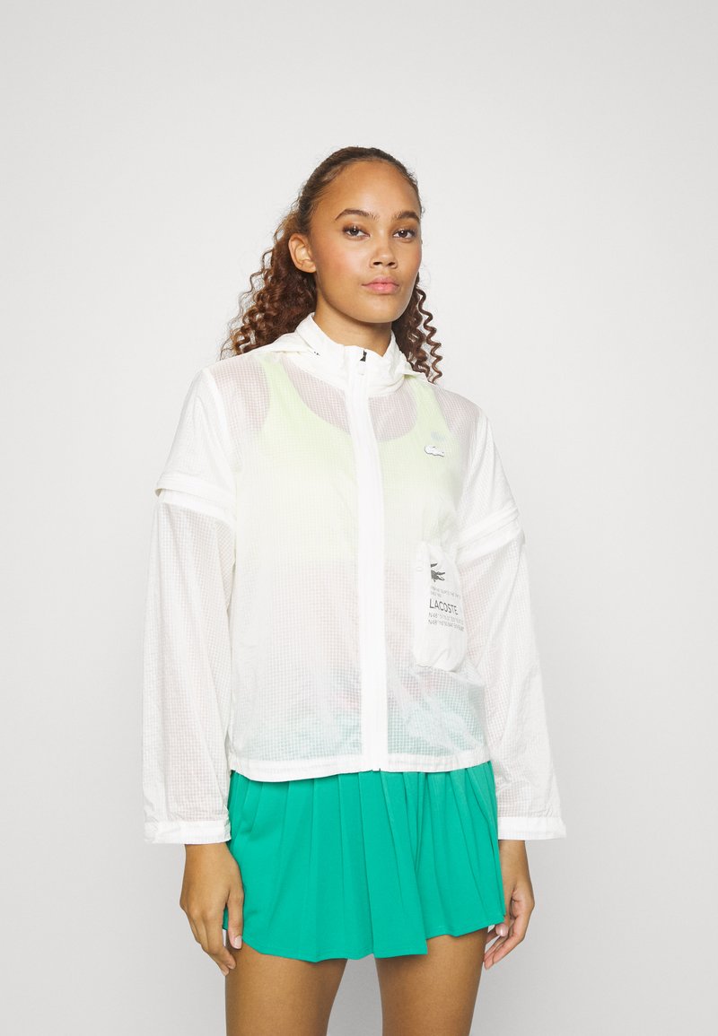 Veste en maille blanche avec capuche, fermeture éclair à l'avant et poche à rabat ; associée à une jupe plissée teal. Tissu léger et respirant.