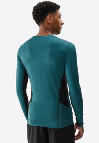 Camisa deportiva de manga larga en azul verdoso con detalles en negro. El material parece suave, presenta un diseño ajustado y bolsillos laterales.