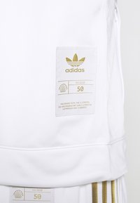 Witte sporttop met een gestructureerde afwerking, voorzien van een goudkleurige Adidas-logopatch met merkinformatie en een maatlabel aan de voorkant.