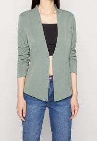 Ljusgrön, figursydd kavaj med långa ärmar, rynkade detaljer och öppen front. Bärs över en svart crop top och kombineras med blå jeans.