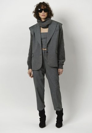 Grijze op maat gemaakte pantalonsuit met een getailleerde blazer met oversized schouders, een gestructureerde wollen sjaal en slanke zwarte enkellaarzen.