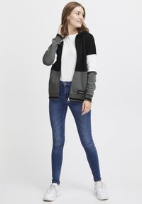 Svart, grå og hvit zip-up jakke med lange ermer, parret med blå jeans og hvite joggesko. Har et logo-patch på jakken.