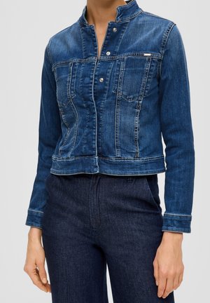 Person trägt eine taillierte blaue Jeansjacke mit Druckknöpfen und hohem Kragen, kombiniert mit dunkelblauer Jeanshose vor einfarbigem Hintergrund.