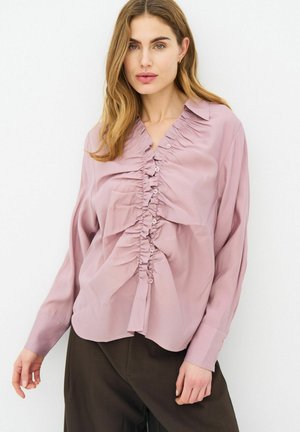 Femme portant un chemisier froncé violet clair à manches longues et un pantalon marron foncé, debout devant un fond blanc uni.