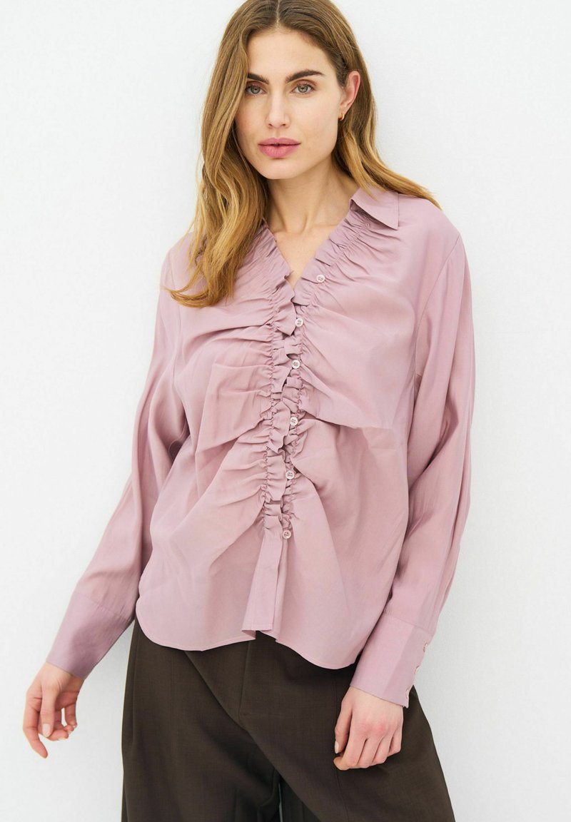Femme portant un chemisier froncé violet clair à manches longues et un pantalon marron foncé, debout devant un fond blanc uni.