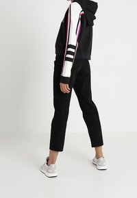 Person wearing black cropped pants, white and black hoodie with pink stripes, and white sneakers standing on white floor against white wall.